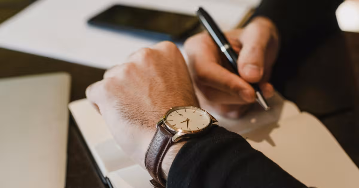 Person checking a wristwatch while writing in a notebook for time tracking